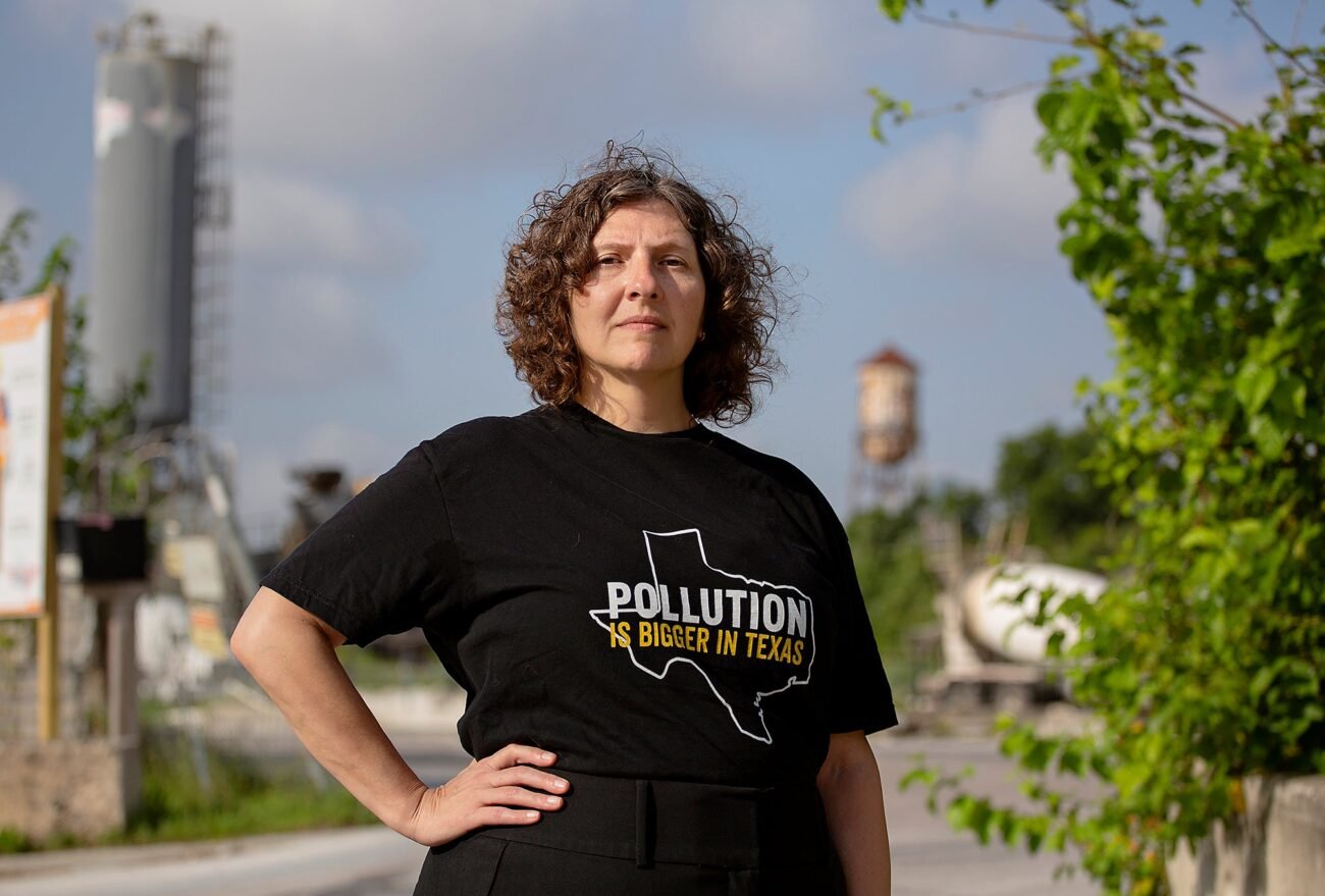 A woman standing next to an industrial facility with a hand on her hip. She is wearing a black t-shirt with an outline of Texas with the words "Pollution is Bigger in Texas"